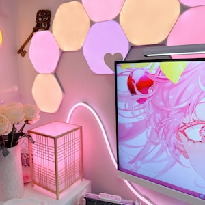 May include: A pink-themed room with hexagonal LED lights, a decorative key, and a square lamp with a grid design. A computer monitor displays a pink anime graphic. A white vase with white roses sits on a white surface.