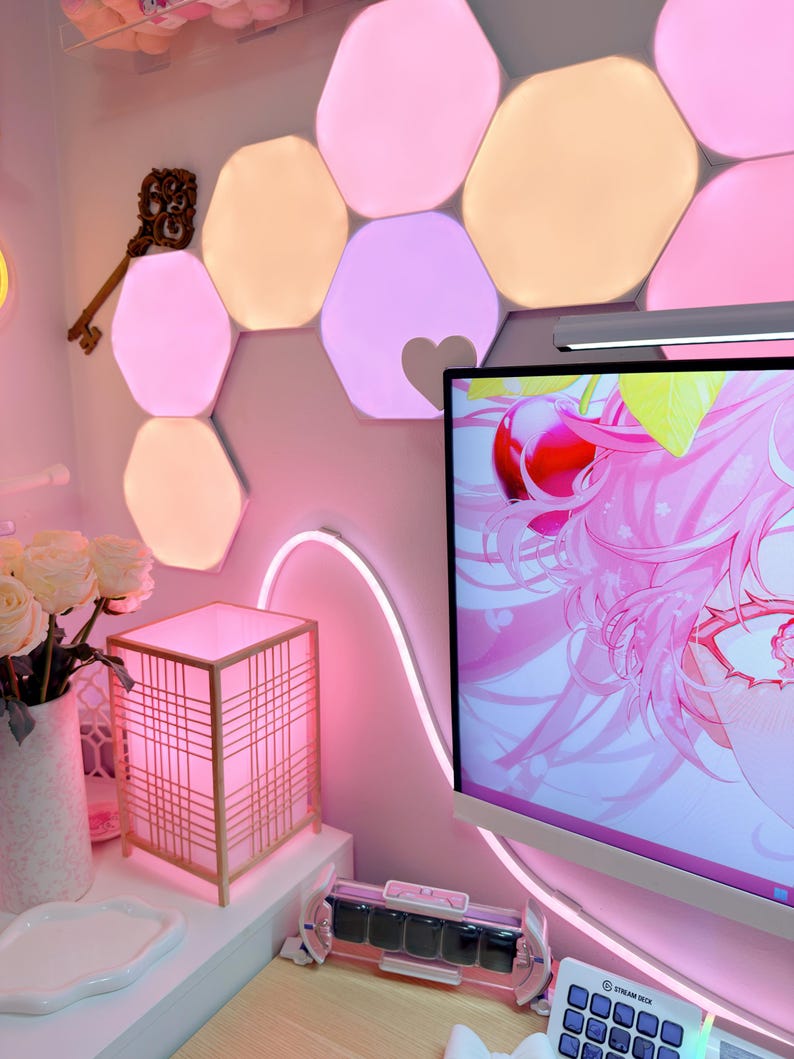 May include: A pink-themed room with hexagonal LED lights, a decorative key, and a square lamp with a grid design. A computer monitor displays a pink anime graphic. A white vase with white roses sits on a white surface.