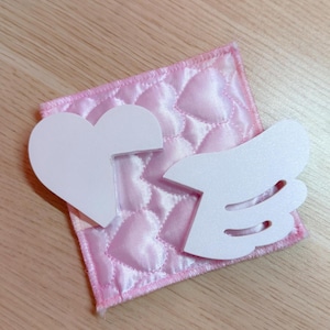 May include: A pink, quilted fabric square with a heart pattern, topped with a white heart and wing-shaped cutouts. The fabric has a satin-like sheen, and the edges are neatly stitched.