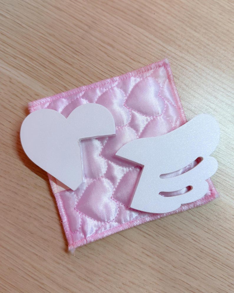 May include: A pink, quilted fabric square with a heart pattern, topped with a white heart and wing-shaped cutouts. The fabric has a satin-like sheen, and the edges are neatly stitched.