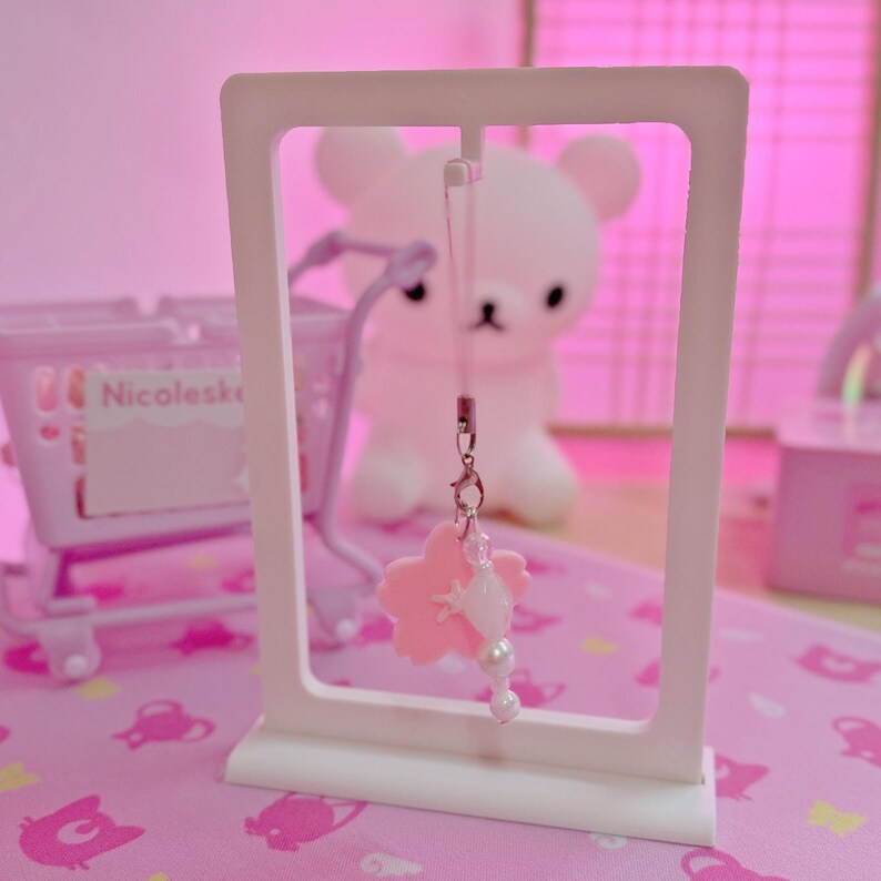 May include: A pink cherry blossom charm with white pearl accents hangs from a white rectangular frame. The charm is suspended in front of a soft pink background with a blurred white bear figure.
