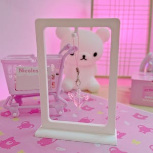May include: A pink display stand holding a clear, winged charm. A miniature shopping trolley and a white bear-shaped light are visible in the background. The scene is set on a pink surface with a repeating cat pattern.