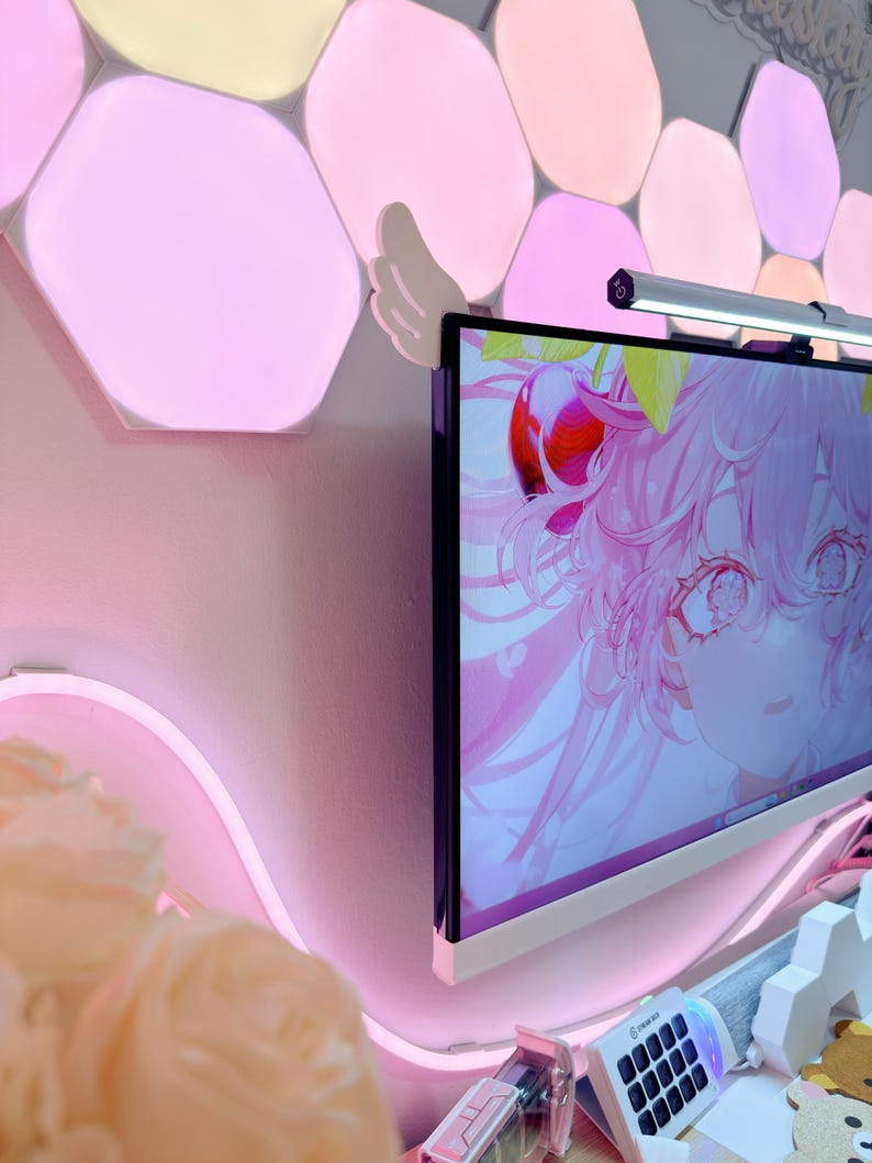May include: A computer monitor displays a pink anime-style graphic. The monitor is mounted on a wall with pink hexagon-shaped lights. A pink neon light and a small keyboard are visible.