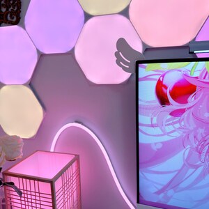 May include: A room with a pink and white aesthetic. Hexagon-shaped LED lights in various shades of pink and white are mounted on the wall. A pink neon light curves along the wall. A pink lamp with a wooden frame sits on a white surface.