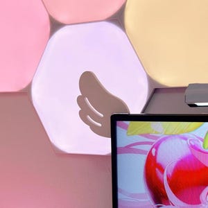 May include: A wall-mounted, hexagonal light panel arrangement in shades of pink and yellow. A light-coloured wing-shaped decoration is attached to one of the panels. A computer monitor displays a colourful image of a cherry.