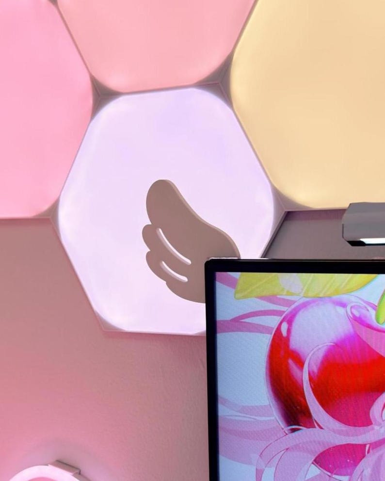 May include: A wall-mounted, hexagonal light panel arrangement in shades of pink and yellow. A light-colored wing-shaped decoration is attached to one of the panels. A computer monitor displays a colorful image of a cherry.
