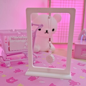 May include: A white rectangular display stand with a small pink shopping cart and a bear-shaped lamp in the background. A charm with a white candy design hangs from the stand. The text "Nicoleskey" is visible on the cart.