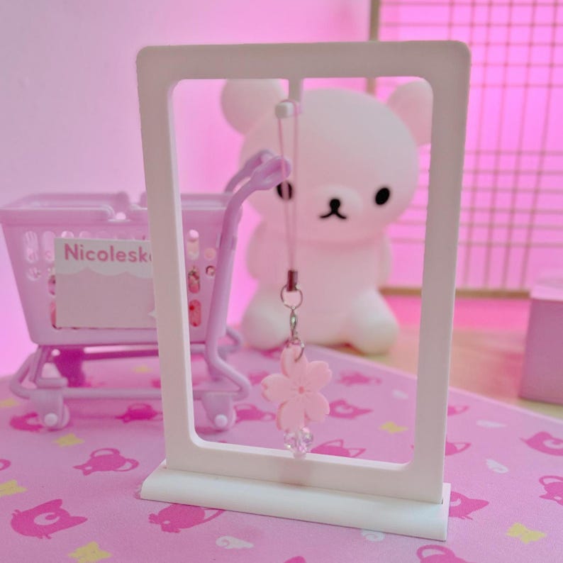 May include: A white display stand showcases a pink cherry blossom charm with a clear crystal bead. A pink shopping trolley and a white bear-shaped lamp are in the background. The scene is set on a pink surface with a matching pink backdrop.