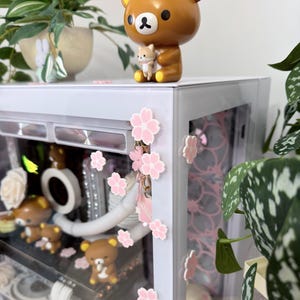 Könnte beinhalten: Eine braune Bärenfigur, die eine kleinere Bärenfigur hält, auf einem weißen Schrank. Rosa Kirschblütendekorationen schmücken die Glastür des Schranks. Weitere Bärenfiguren und dekorative Elemente sind im Inneren des Schranks zu sehen.
