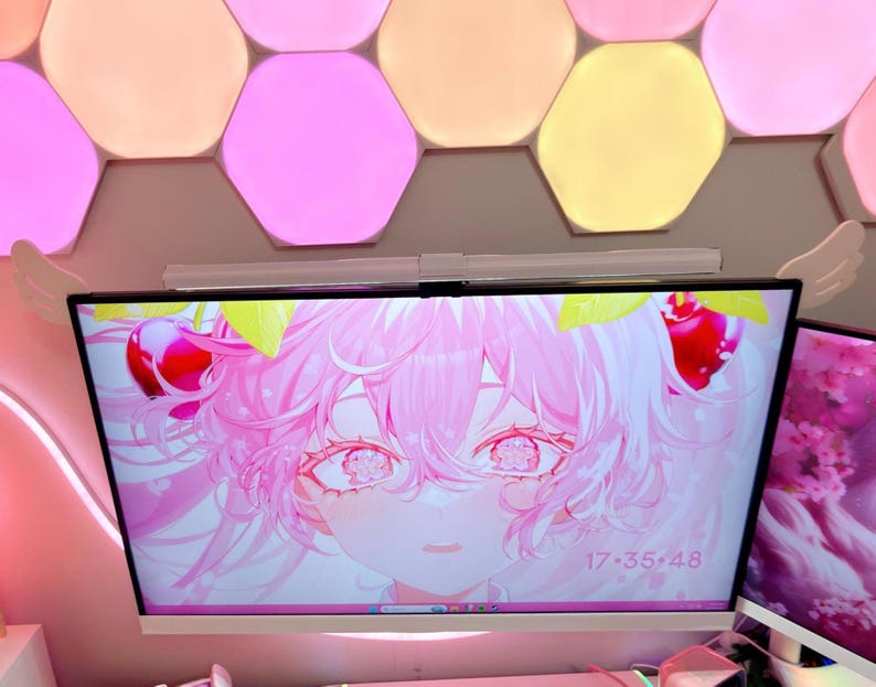 May include: A computer monitor displays a pink anime-style character with cherry accents and the time 17:35:48. The monitor is mounted on a wall with a light fixture and decorative wing accents. Hexagon-shaped lights in pink, yellow, and orange are above.