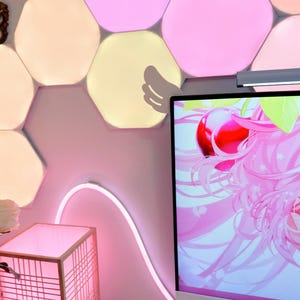 May include: A pink-themed room with a computer monitor displaying a pink anime graphic. A pink neon light curves along the wall. A wooden lantern and hexagon-shaped wall lights add to the decor. The room has a modern, colorful aesthetic.
