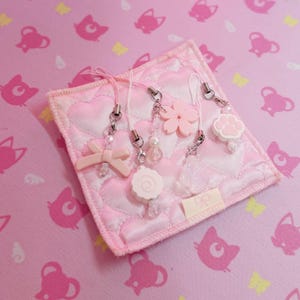 May include: A selection of pink and white charms, including a bow, flower, paw print, and swirl, attached to a pink quilted square. The charms are on white cords with silver clasps. The background is a pink fabric with cat and butterfly patterns.