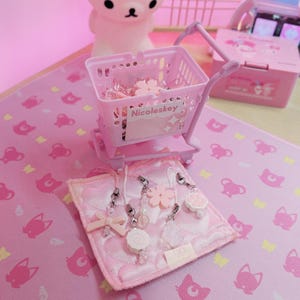 May include: A pink miniature shopping trolley filled with charms and accessories, with the text "Nicoleskey" on the side. The charms include flowers, bows, and other decorative elements. The items are displayed on a pink surface.
