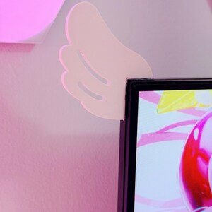 May include: A light pink, wing-shaped wall decor piece is mounted on a pink wall, positioned near the corner of a black computer monitor. The wing has a layered design with three visible sections.