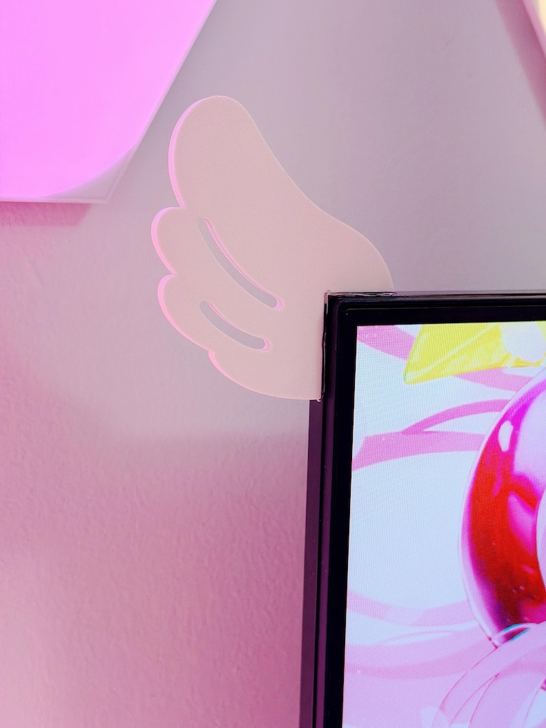 May include: A light pink, wing-shaped wall decor piece is mounted on a pink wall, positioned near the corner of a black computer monitor. The wing has a layered design with three visible sections.