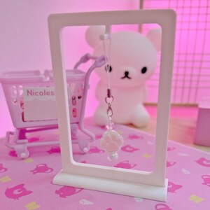 May include: A pink paw print charm with a white pearl hangs in front of a white rectangular frame. A pink shopping trolley and a white bear figurine are in the background. The background is pink.