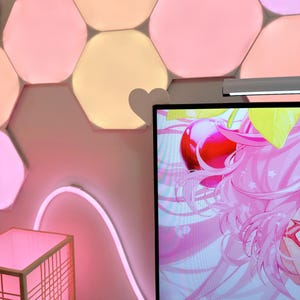 May include: A room with pink and peach-colored hexagon-shaped wall lights. A pink neon light curves along the wall. A monitor displays a pink anime graphic. A small, square, pink-lit lamp is in the foreground.