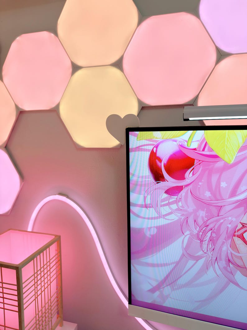 May include: A room with pink and peach-colored hexagon-shaped wall lights. A pink neon light curves along the wall. A monitor displays a pink anime graphic. A small, square, pink-lit lamp is in the foreground.
