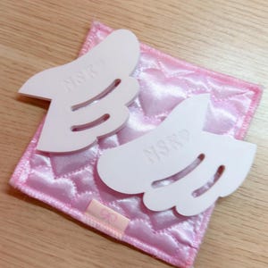 May include: Two white, wing-shaped objects with the letters "NSKO" are placed on a pink, quilted square. The square is on a light brown wooden surface. The wings have curved cutouts and are slightly raised.