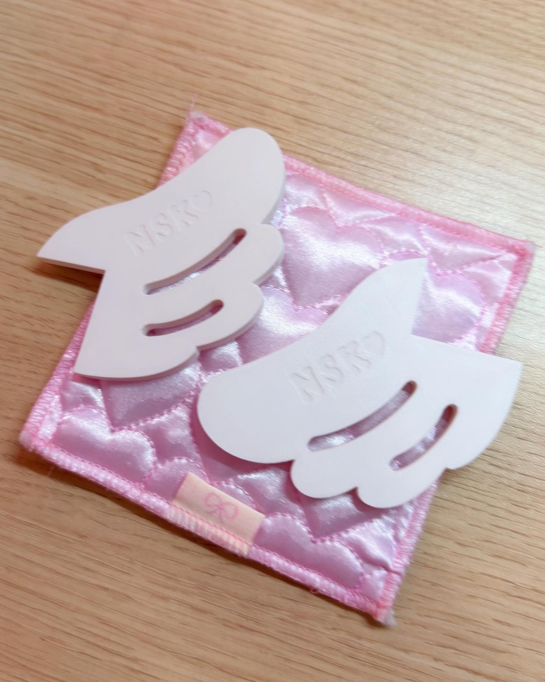 May include: Two white, wing-shaped objects with the letters "NSKO" are placed on a pink, quilted square. The square is on a light brown wooden surface. The wings have curved cutouts and are slightly raised.