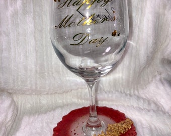 May include: A clear glass wine glass with a gold inscription that reads "Happy Mother's Day". The glass is sitting on a red and white resin coaster with gold glitter.