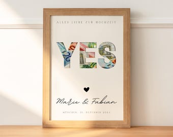 May include: A framed print with a white background and the word "YES" in large letters. The letters are made of cut-out pieces of paper money. Below the word "YES" is a small black heart and the text "Marie & Fabian" in cursive. The text "Mönchen, 31. Dezember 2024" is printed below the names. The frame is a light brown wood.