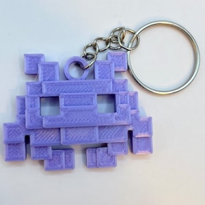 Könnte beinhalten: Ein lilafarbener, 3D-gedruckter Schlüsselanhänger in Form eines pixeligen Space Invaders.