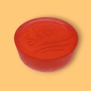 Peut inclure: Un savon rond rouge avec le texte en relief "Marie Savons" et des lignes ondulées. Le savon a un aspect lisse et légèrement translucide, sur un fond orange clair.