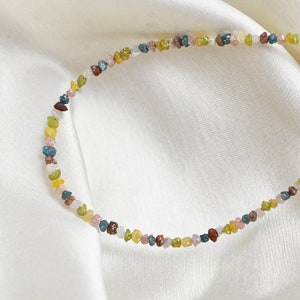 May include: A delicate necklace made of small, colorful, rough-cut gemstones. The necklace is made of a variety of colors, including pink, green, blue, and yellow.