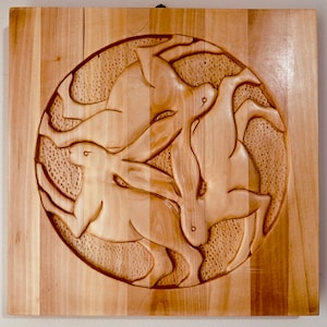 May include: A wooden wall sculpture of three hares carved in a circle. The hares are in a continuous loop, with their tails and heads touching. The sculpture is made of light brown wood and has a natural finish.