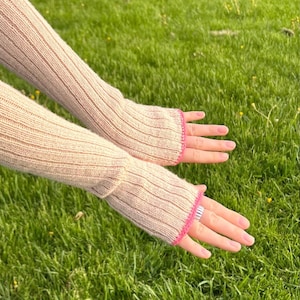 Könnte beinhalten: Beige gestrickte Armstulpen mit Rippenmuster und rosa Einfassung. Die Armstulpen werden vor einem Hintergrund aus grünem Gras gezeigt.