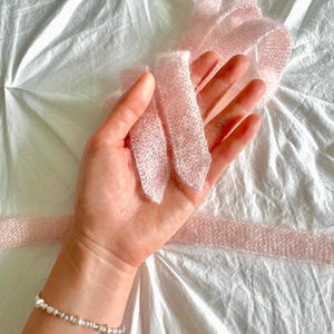 May include: A delicate, light pink knitted ribbon is held in a hand, with a pearl and silver bead bracelet on the wrist. The ribbon is soft and textured, with a slightly fuzzy appearance. The background is a white, textured fabric.