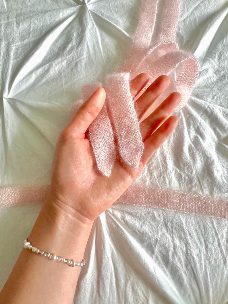 May include: A delicate, light pink knitted ribbon is held in a hand, with a pearl and silver bead bracelet on the wrist. The ribbon is soft and textured, with a slightly fuzzy appearance. The background is a white, textured fabric.