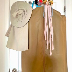 May include: A tan garment bag hangs on a white door, with a wide-brimmed hat and a decorative floral accessory. The hat is light beige, and the garment bag is a medium brown. A colorful, flower-shaped accessory with pink streamers is attached to the top.