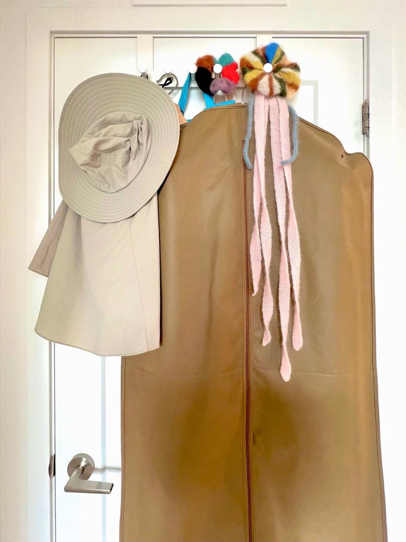 May include: A tan garment bag hangs on a white door, with a wide-brimmed hat and a decorative floral accessory. The hat is light beige, and the garment bag is a medium brown. A colorful, flower-shaped accessory with pink streamers is attached to the top.
