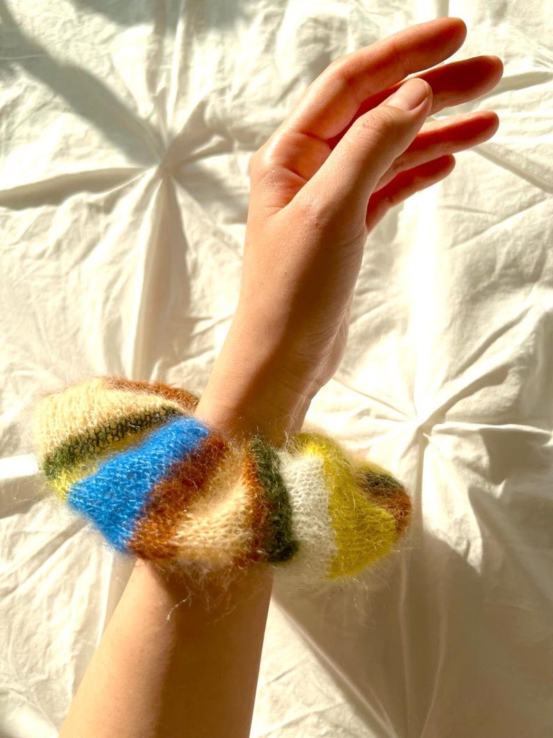 May include: A colorful scrunchie with a mix of blue, yellow, green, brown, and white stripes. The scrunchie is made of a soft, textured material and is worn on the wrist. The scrunchie is a hair accessory.