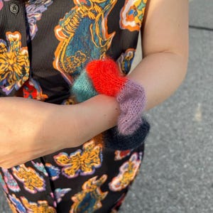 May include: A person wearing a black patterned top and pants holds a collection of colorful, fuzzy, knitted wrist warmers. The wrist warmers are in shades of red, teal, purple, and navy blue. The top and pants have a repeating pattern of colorful, stylized fish.