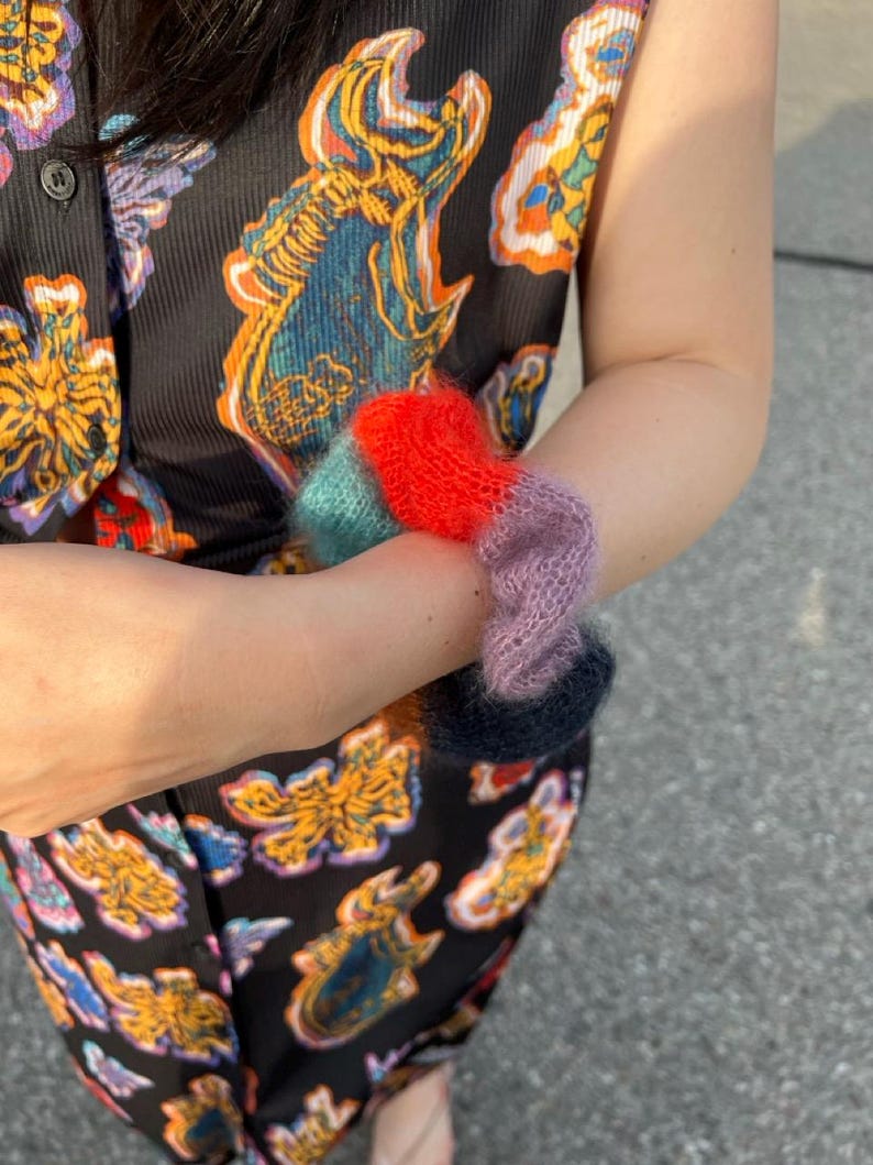 May include: A person wearing a black patterned top and pants holds a collection of colorful, fuzzy, knitted wrist warmers. The wrist warmers are in shades of red, teal, purple, and navy blue. The top and pants have a repeating pattern of colorful, stylized fish.