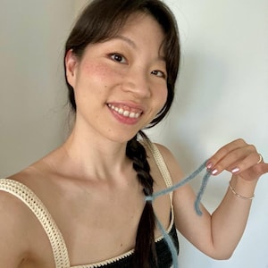 May include: A black crochet top with cream-colored straps and trim. A strand of light blue yarn is held up. The top is handmade and appears to be part of a set. The image is well-lit and shows the texture of the crochet.