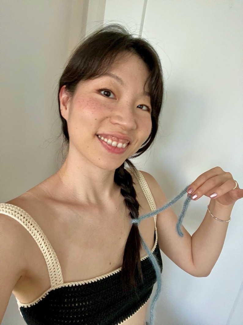 May include: A black crochet top with cream-colored straps and trim. A strand of light blue yarn is held up. The top is handmade and appears to be part of a set. The image is well-lit and shows the texture of the crochet.