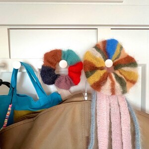May include: Two colorful, fluffy, knitted accessories hang on a white door. One is a multi-colored donut shape, and the other is a flower shape with pink and blue tassels. A blue tote bag and a tan jacket are also visible.