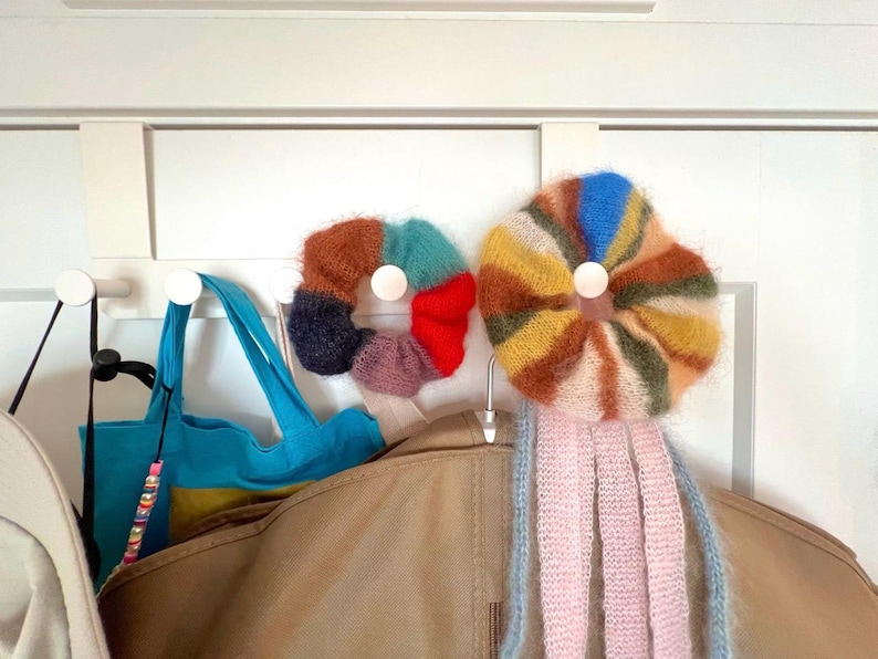 May include: Two colorful, fluffy, knitted accessories hang on a white door. One is a multi-colored donut shape, and the other is a flower shape with pink and blue tassels. A blue tote bag and a tan jacket are also visible.