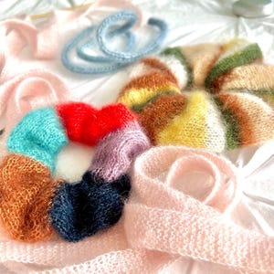 May include: A collection of handmade, fluffy hair scrunchies and a scarf in various colors. The scrunchies are in shades of red, blue, orange, and purple. A light pink scarf and a light blue hair tie are also visible.