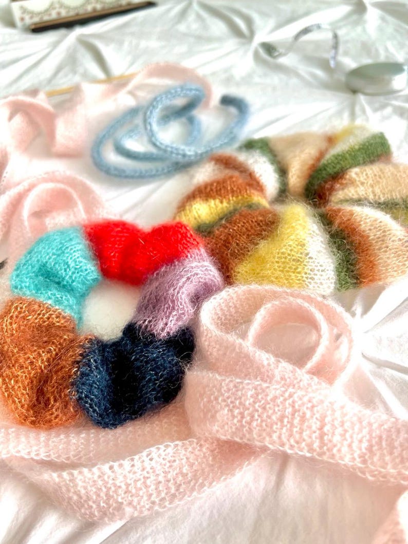 May include: A collection of handmade, fluffy hair scrunchies and a scarf in various colors. The scrunchies are in shades of red, blue, orange, and purple. A light pink scarf and a light blue hair tie are also visible.