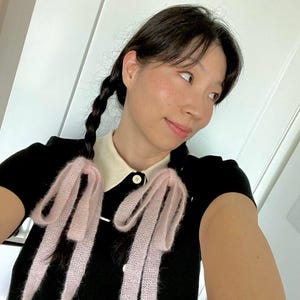 May include: A black short-sleeved top with a white collar and white trim. The top features two light pink, fluffy bows. The buttons are white. The person has dark hair in braids.