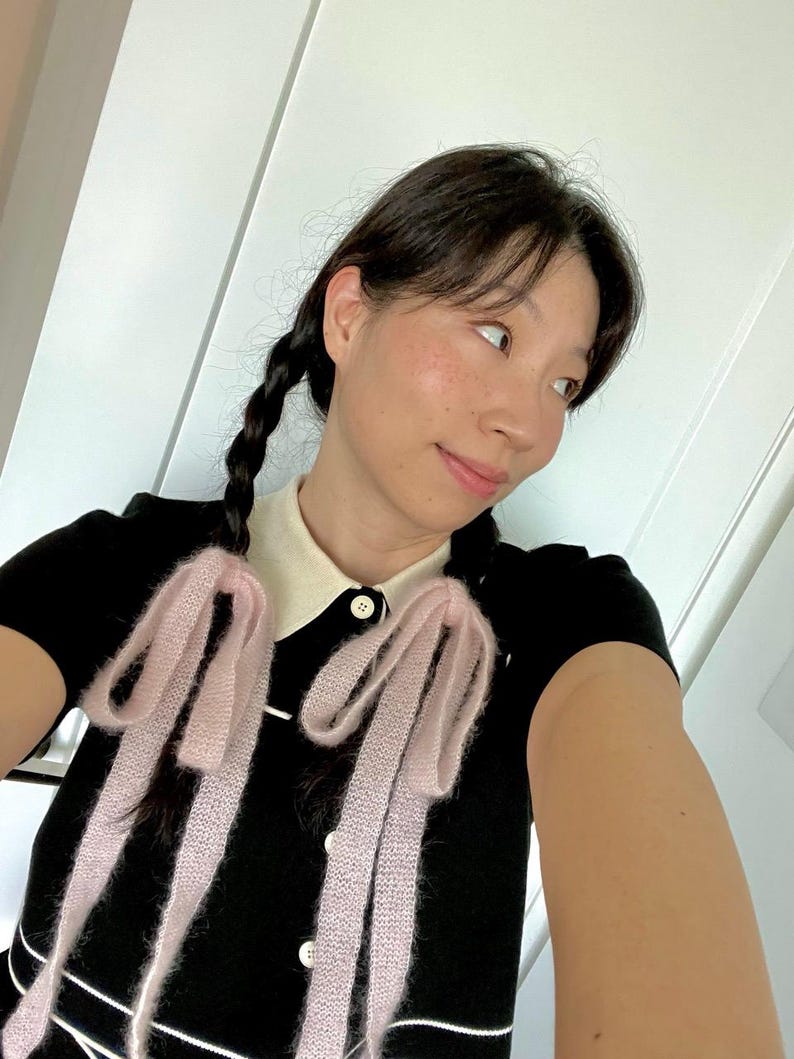 May include: A black short-sleeved top with a white collar and white trim. The top features two light pink, fluffy bows. The buttons are white. The person has dark hair in braids.