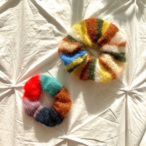 May include: Two colourful knitted scrunchies. One is a mix of red, orange, teal, and purple. The other is striped with yellow, brown, green, and blue. Both are on a white, textured surface.