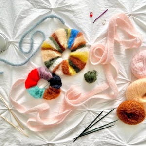 May include: An assortment of colorful yarn and knitting supplies are arranged on a white surface. The items include two knitted scrunchies, yarn balls, knitting needles, and a tape measure. The scrunchies feature a variety of colors.