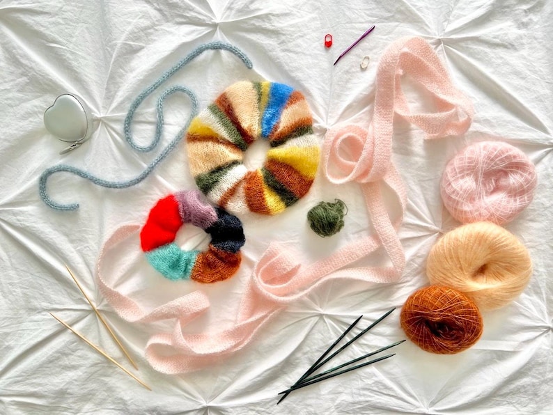 May include: An assortment of colorful yarn and knitting supplies are arranged on a white surface. The items include two knitted scrunchies, yarn balls, knitting needles, and a tape measure. The scrunchies feature a variety of colors.