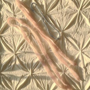 May include: Two pairs of light pink, fuzzy leg warmers and a silver beaded necklace are arranged on a cream-colored, textured surface. The leg warmers are long and slender, with a soft, textured appearance.